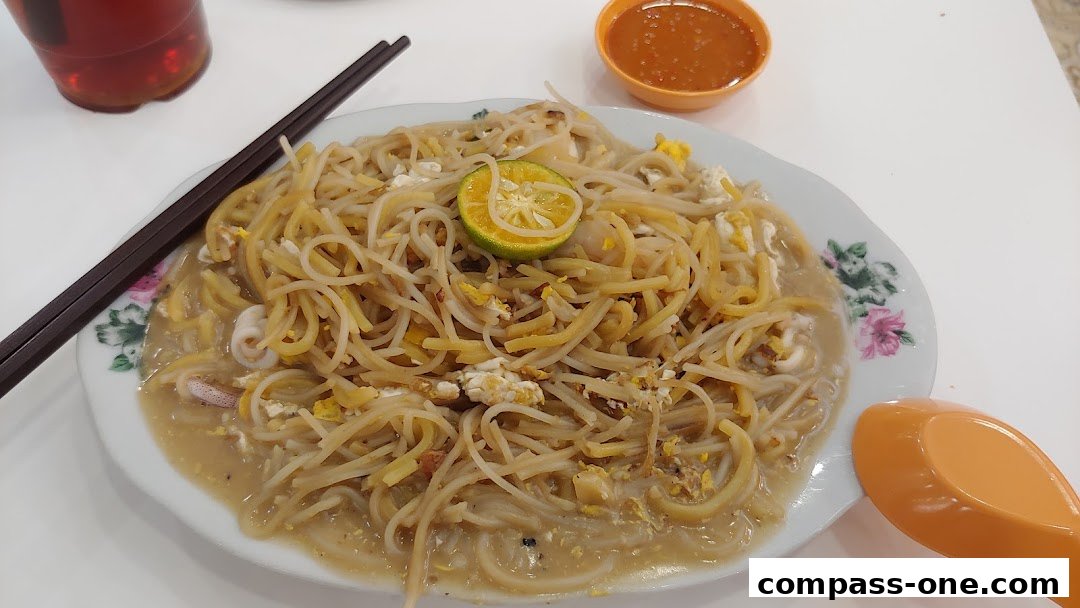 A plate of noodles with squid, egg, and a lime wedge, served with chili sauce and a drink.