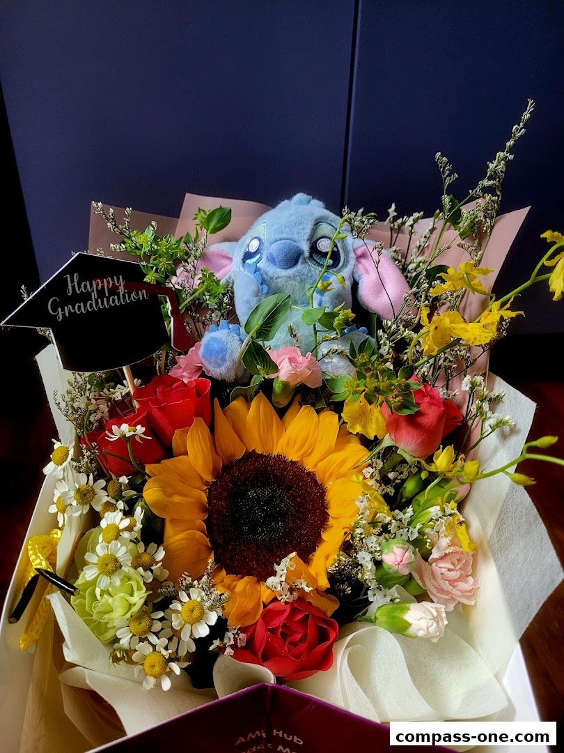 A vibrant bouquet featuring a sunflower, roses, daisies, and a Stitch plush toy with a graduation cap.