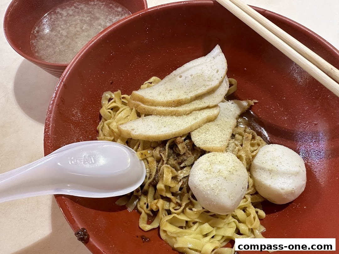 A bowl of noodles with fish cakes, fish slices, and a side of soup, served with chopsticks and a spoon.