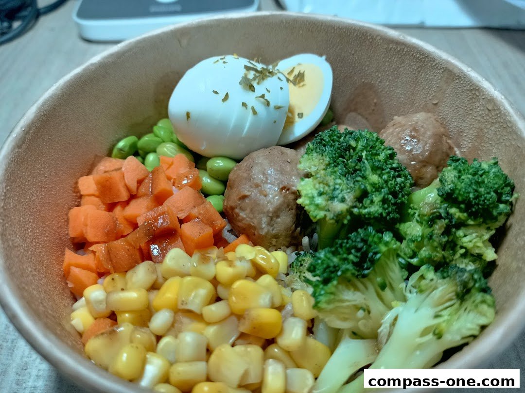 A close-up of a healthy bowl with rice, meatballs, broccoli, carrots, edamame, corn, and a boiled egg.