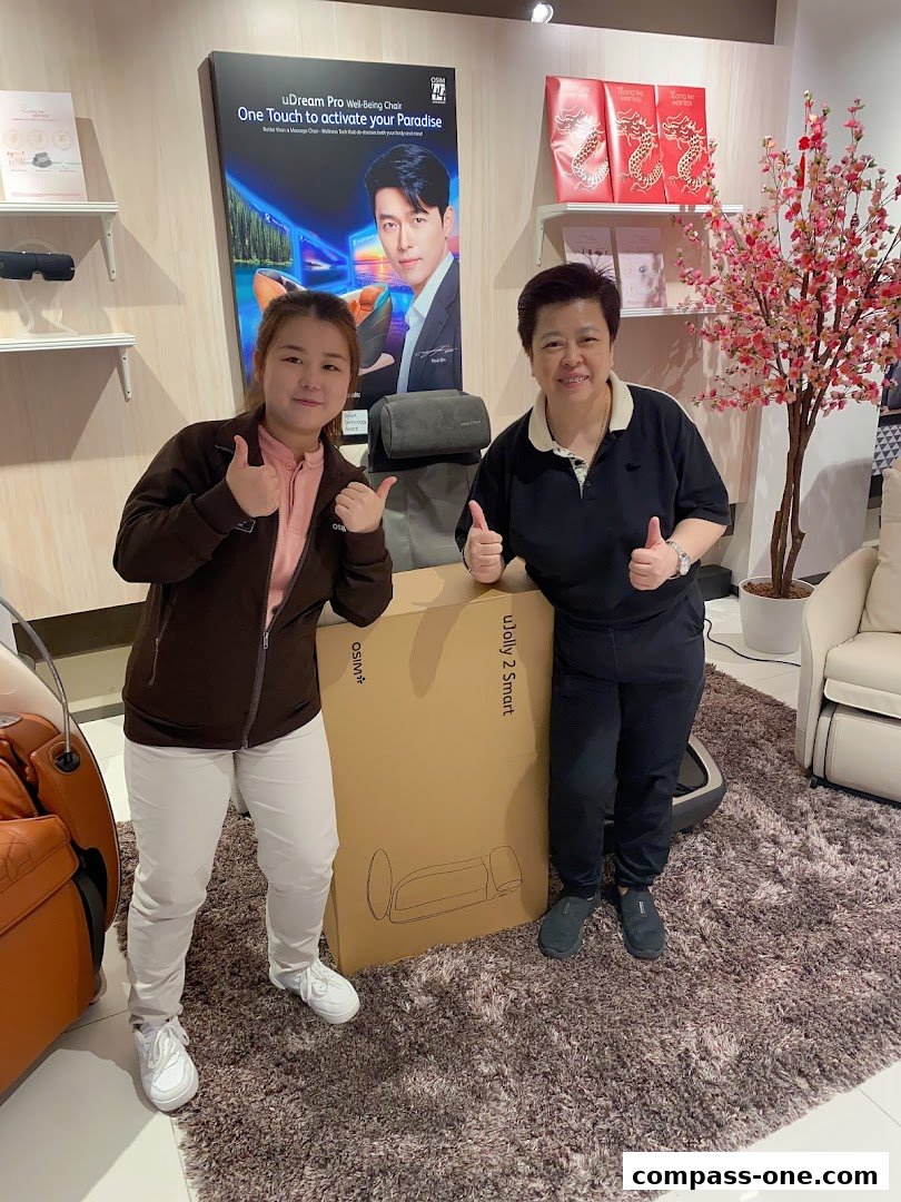 Two smiling women give thumbs up in front of a massage chair box and promotional poster.