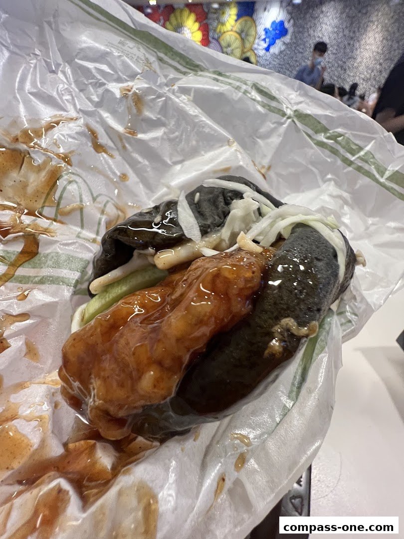 A close-up of a burger with dark buns and a saucy filling, wrapped in paper.