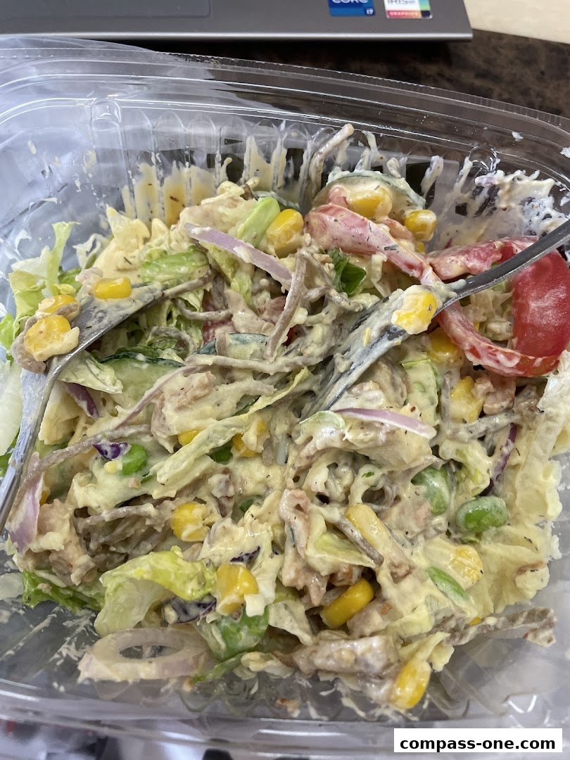A close-up of a salad in a plastic container with forks, featuring lettuce, corn, tomatoes, and a creamy dressing.