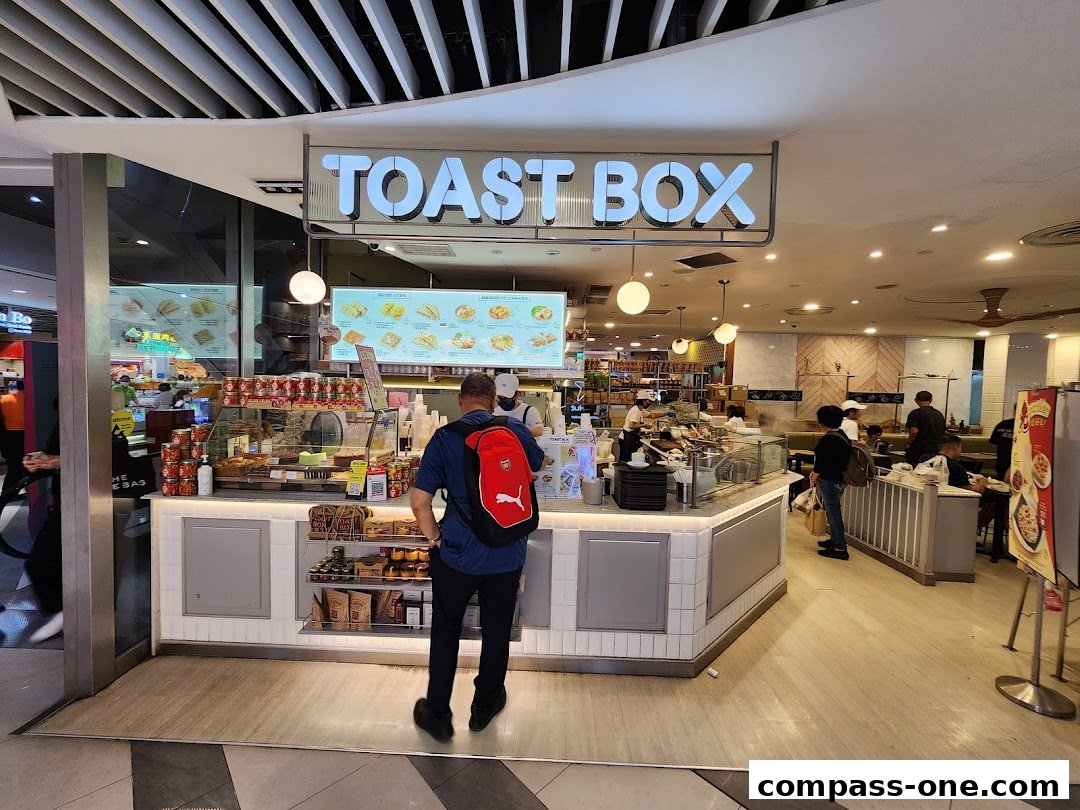 A customer stands at the counter of a Toast Box cafe, with food displays and seating visible.