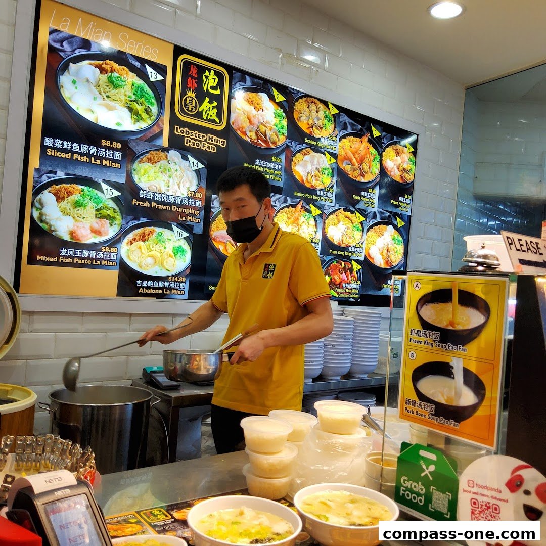 A man in a yellow shirt stands behind a counter with a menu board displaying various noodle dishes.