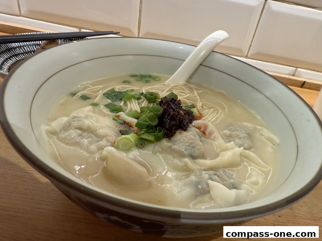 A close-up shot of a steaming bowl of wonton noodle soup with fresh scallions and chili paste.