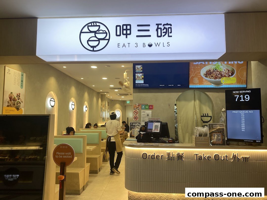 The interior of Eat 3 Bowls restaurant with seating, a counter, and a queue display.
