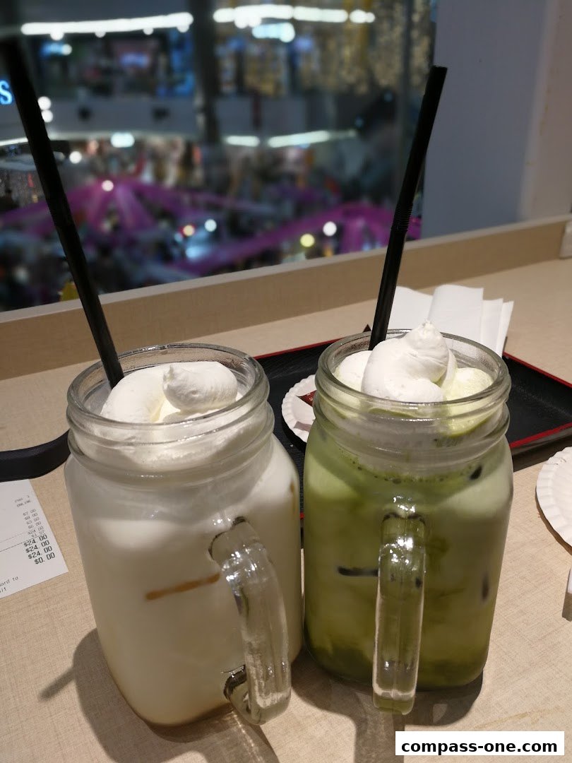 Two iced drinks with whipped cream and straws in mason jars, with a blurred mall interior in the background.