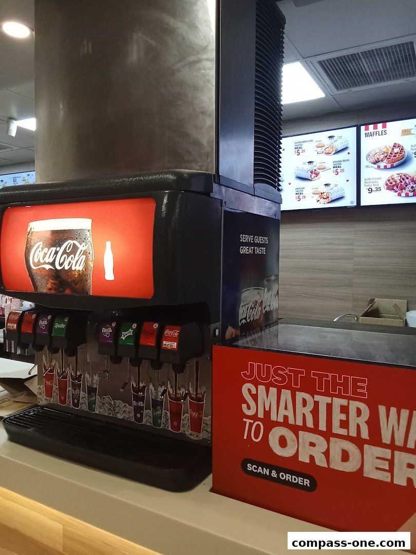 A KFC drinks dispenser with a Coca-Cola advertisement and a menu board in the background.