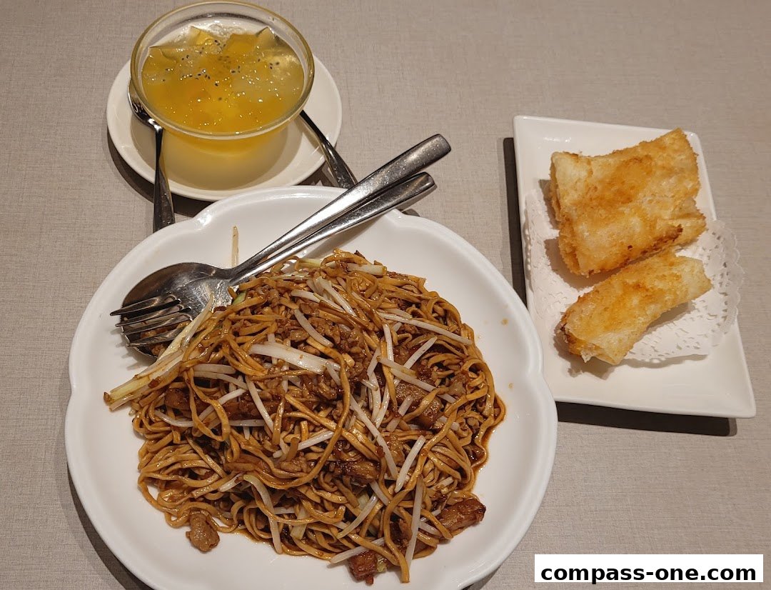 A plate of stir-fried noodles with a side of fried spring rolls and a dessert drink.