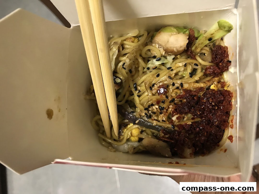 A close-up shot of a takeaway box filled with noodles, chicken, vegetables, and chili paste.
