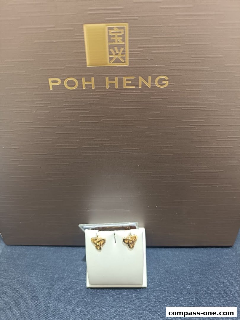 A pair of gold knot-shaped earrings displayed on a white stand in front of a Poh Heng Jewellery branded box.