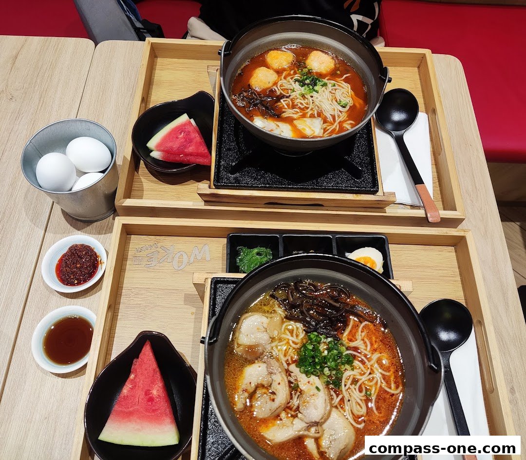 Two steaming bowls of ramen with various toppings, served with watermelon slices and condiments.