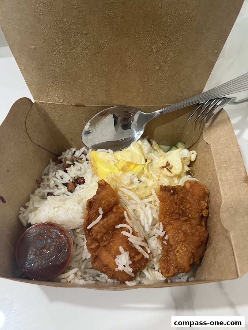 A takeaway box filled with Nasi Lemak, featuring rice, fried chicken, egg, and sambal.