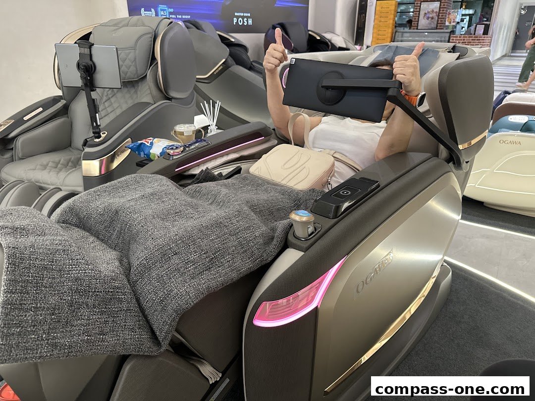 A person enjoys a massage chair with a tablet and snacks, giving a thumbs up.