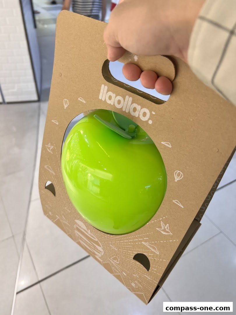 A hand holds a llaollao branded paper bag with a bright green apple-shaped container inside.