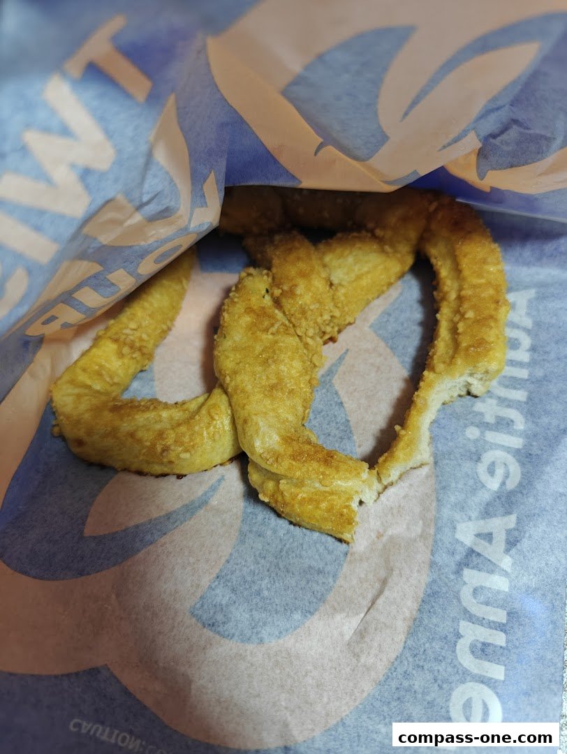 A close-up shot of a golden-brown pretzel with a bite taken out, resting in a branded paper bag.