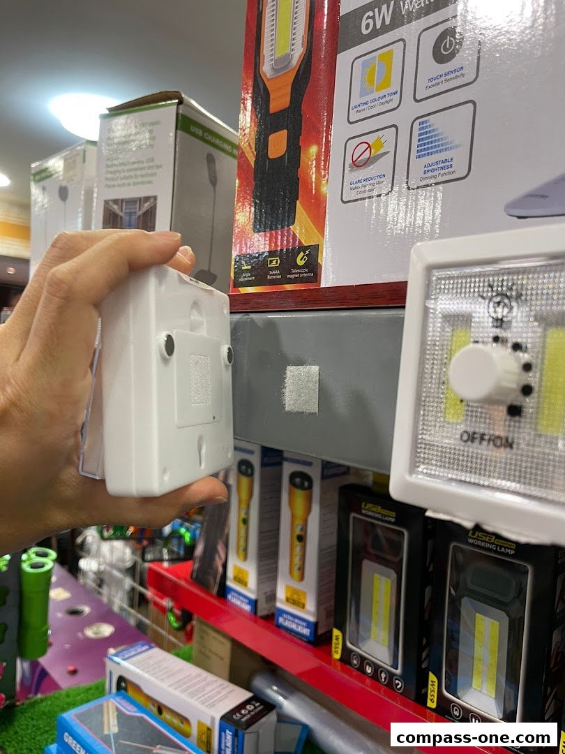 A hand holds a white magnetic light, with various flashlights and work lamps displayed on shelves.
