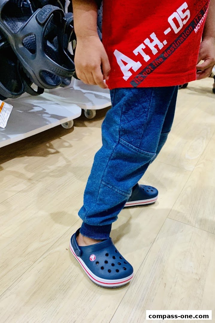 A child wearing blue Crocs and jeans stands in front of a display of black sandals.