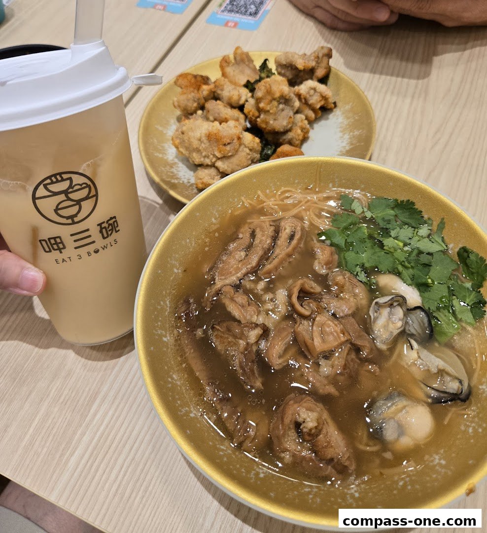 A hand holds a drink next to a bowl of noodle soup and a plate of fried chicken.