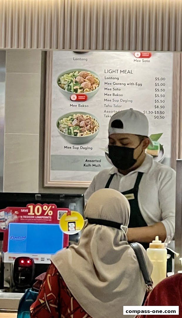 A menu board displays various noodle dishes and light meals with prices, with staff and customers in the foreground.