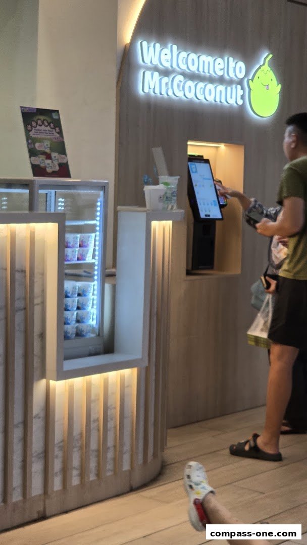 A person orders at the Mr. Coconut counter with a digital menu and refrigerated products.