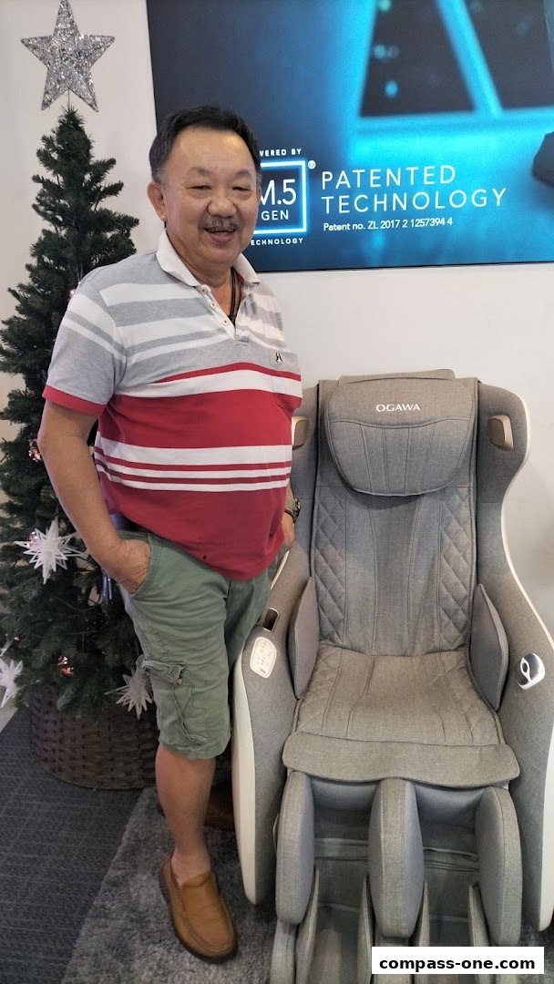 A man stands next to an OGAWA massage chair, with a Christmas tree in the background.