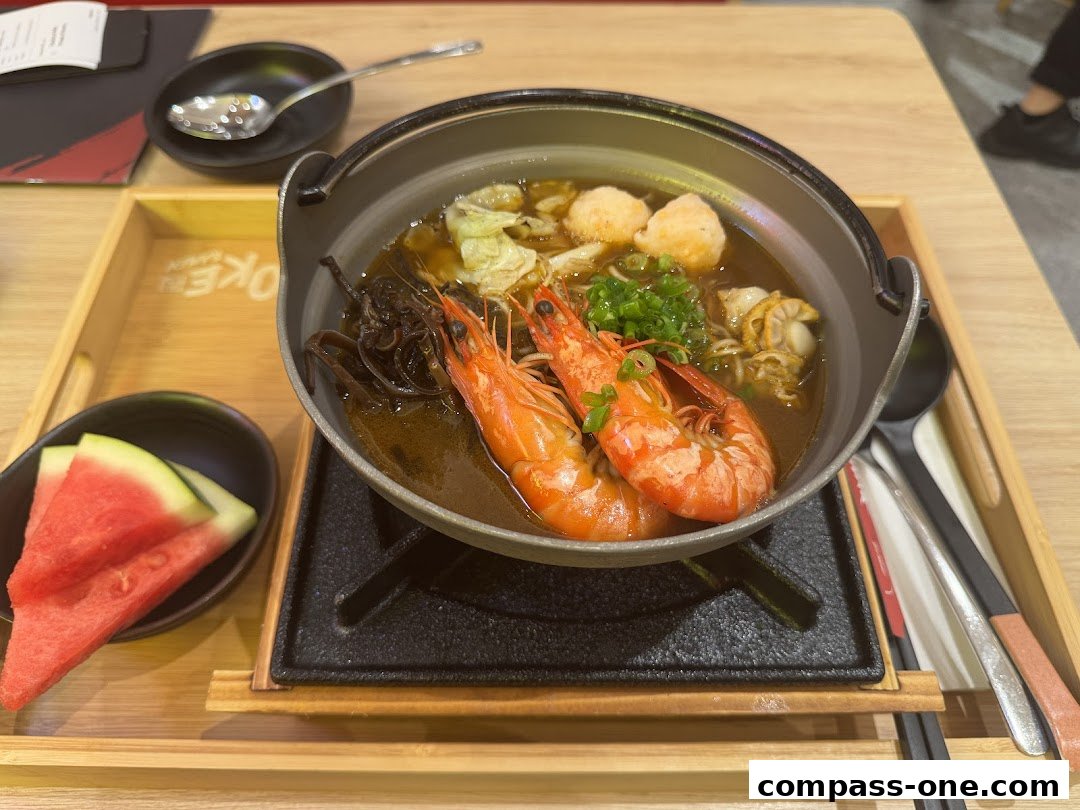 A steaming bowl of ramen with prawns, scallops, and vegetables, served with watermelon slices.