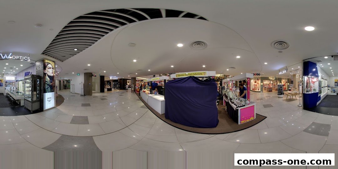 A panoramic view of a shopping mall corridor with various shops, including W Optics.