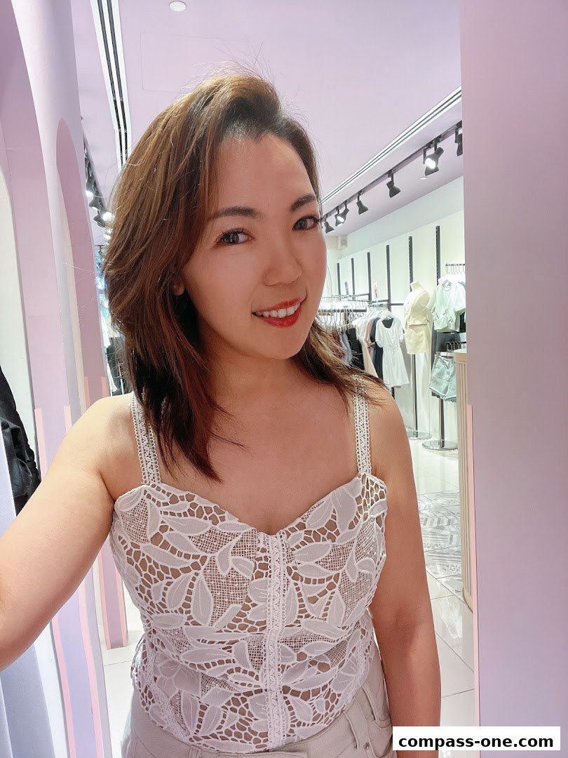 A woman smiles at the camera in a brightly lit clothing store, showcasing a lace top.