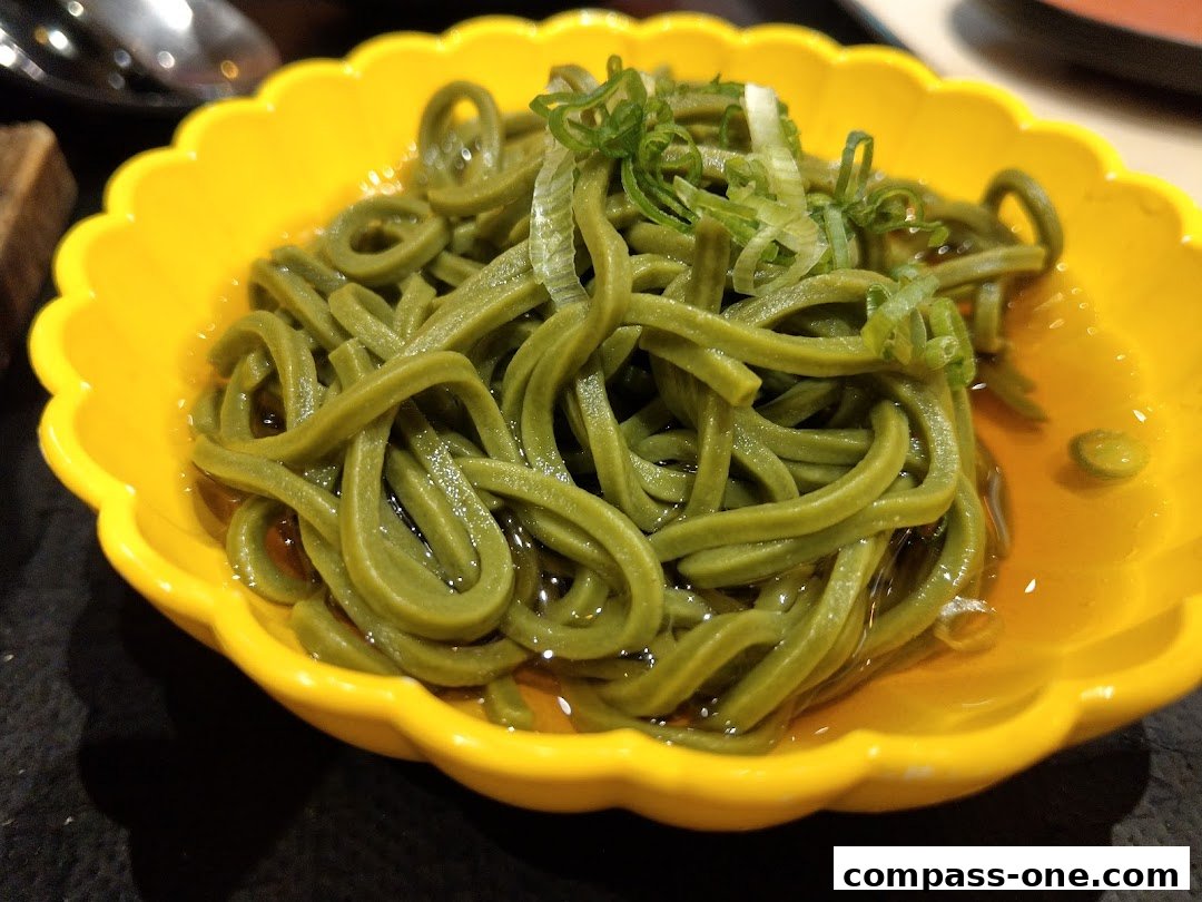 A close-up of green soba noodles served in a bright yellow scalloped bowl with broth and green onions.