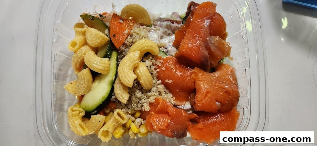 A clear plastic container filled with a colorful salad featuring salmon, pasta, quinoa, zucchini, and carrots.