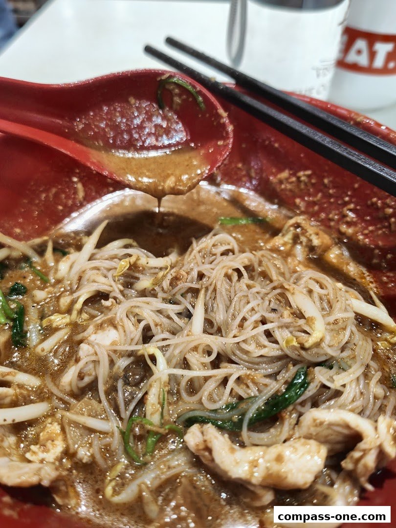 A close-up of a delicious bowl of noodles with chicken and bean sprouts, served with a spoon and chopsticks.