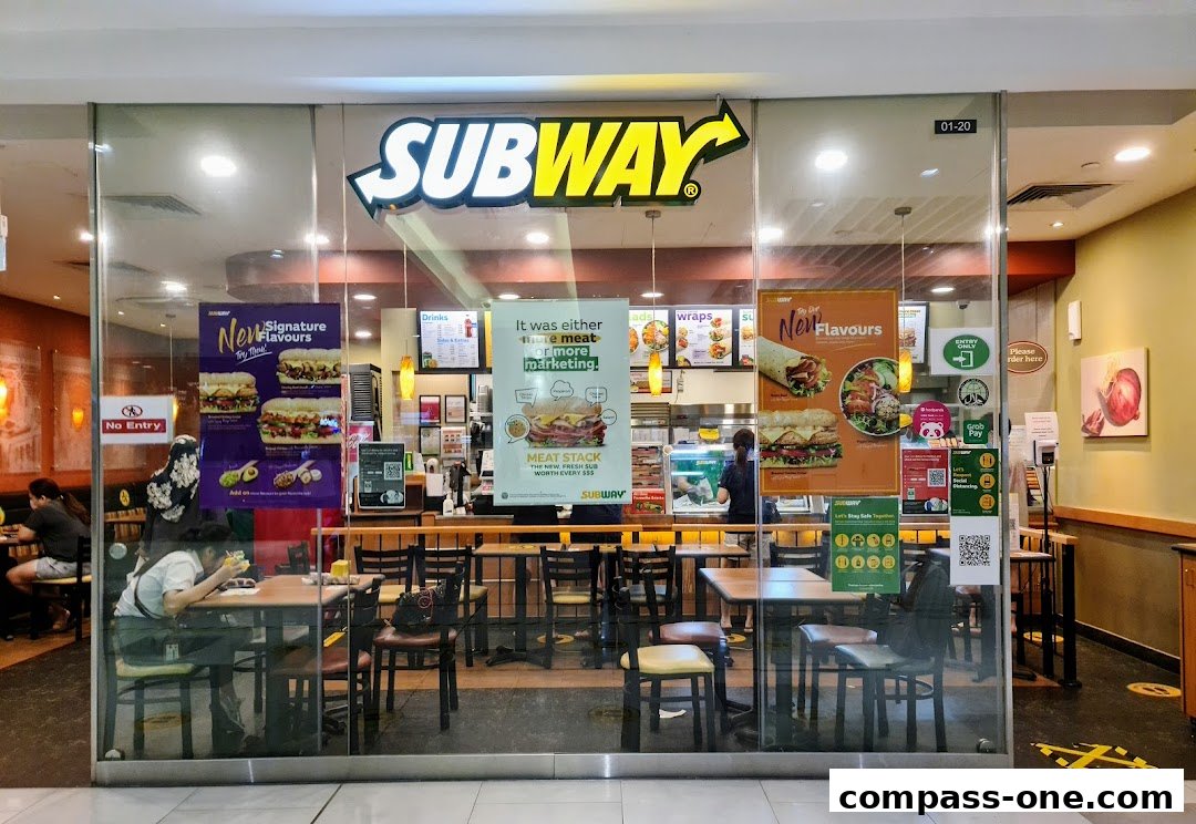 The exterior of a Subway restaurant with customers dining inside and promotional posters.
