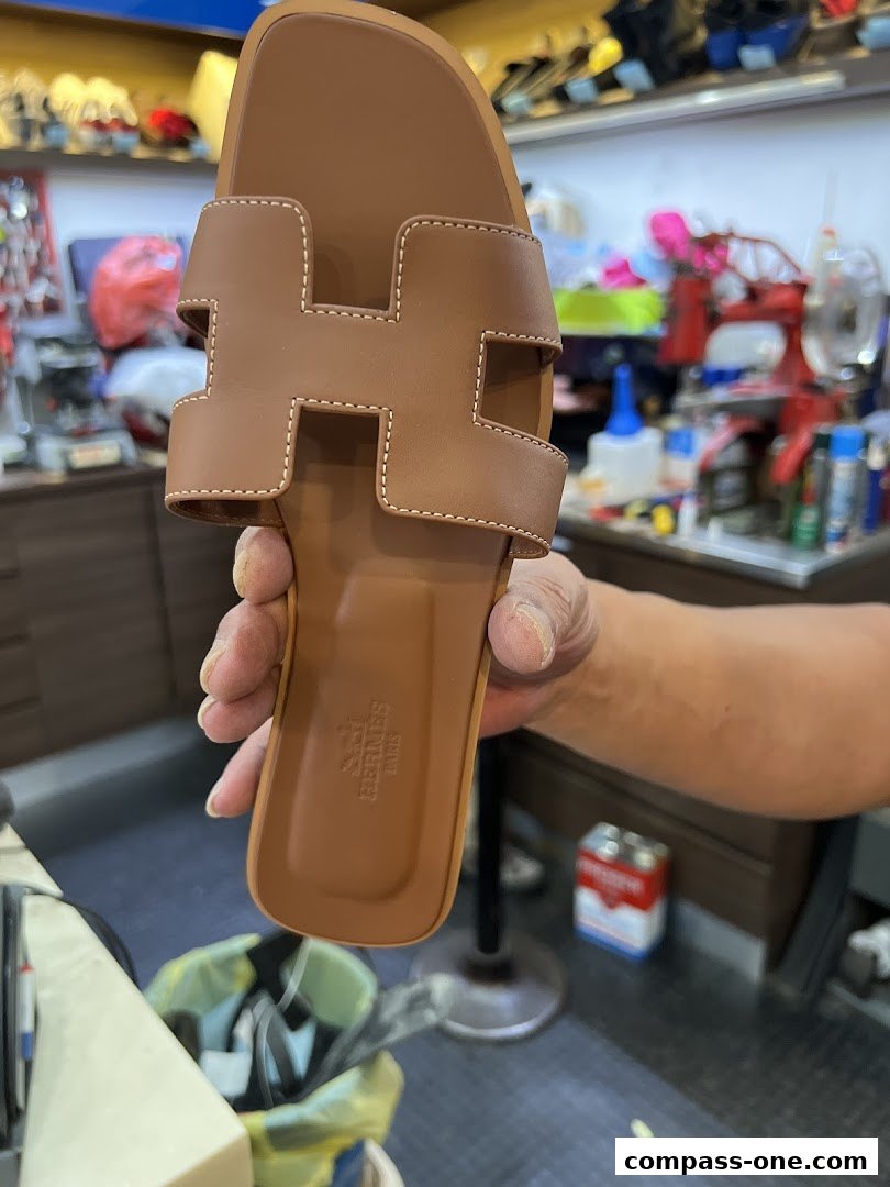 A hand holds a brown sandal with an H design, showing the embossed logo.