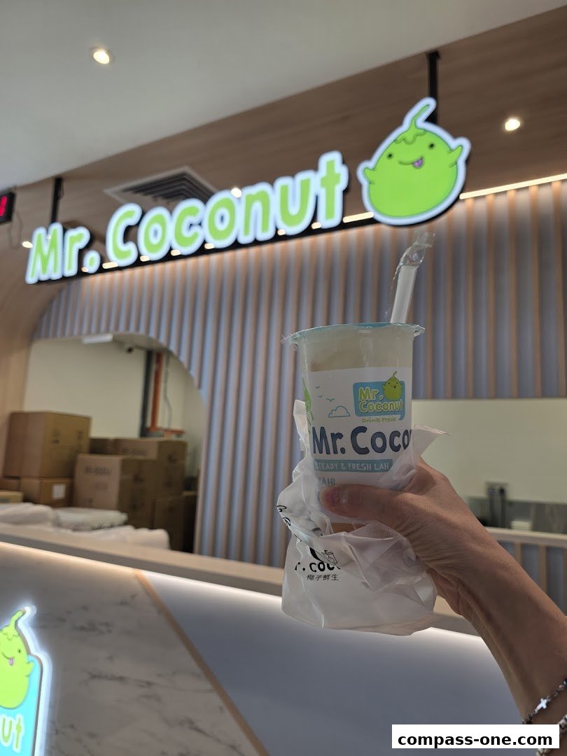 A hand holds a Mr. Coconut drink in front of the shop's illuminated sign.