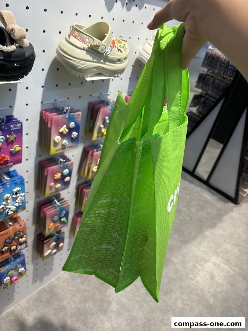 A hand holds a green Crocs branded bag in front of a display of Crocs shoes and Jibbitz.