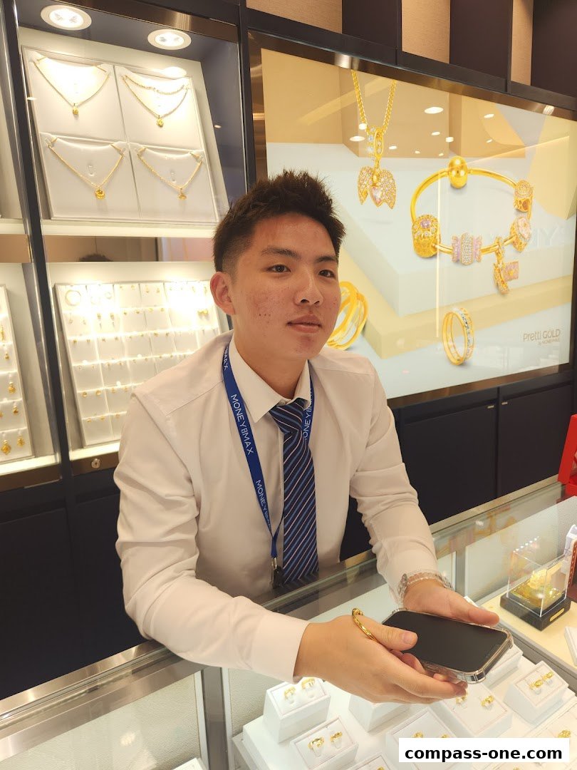 A male staff member stands behind a counter displaying gold jewelry and a promotional poster.