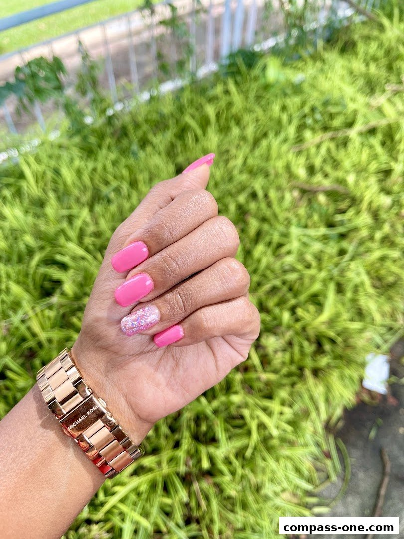 A hand with bright pink nails and a glitter accent nail, wearing a rose gold watch.