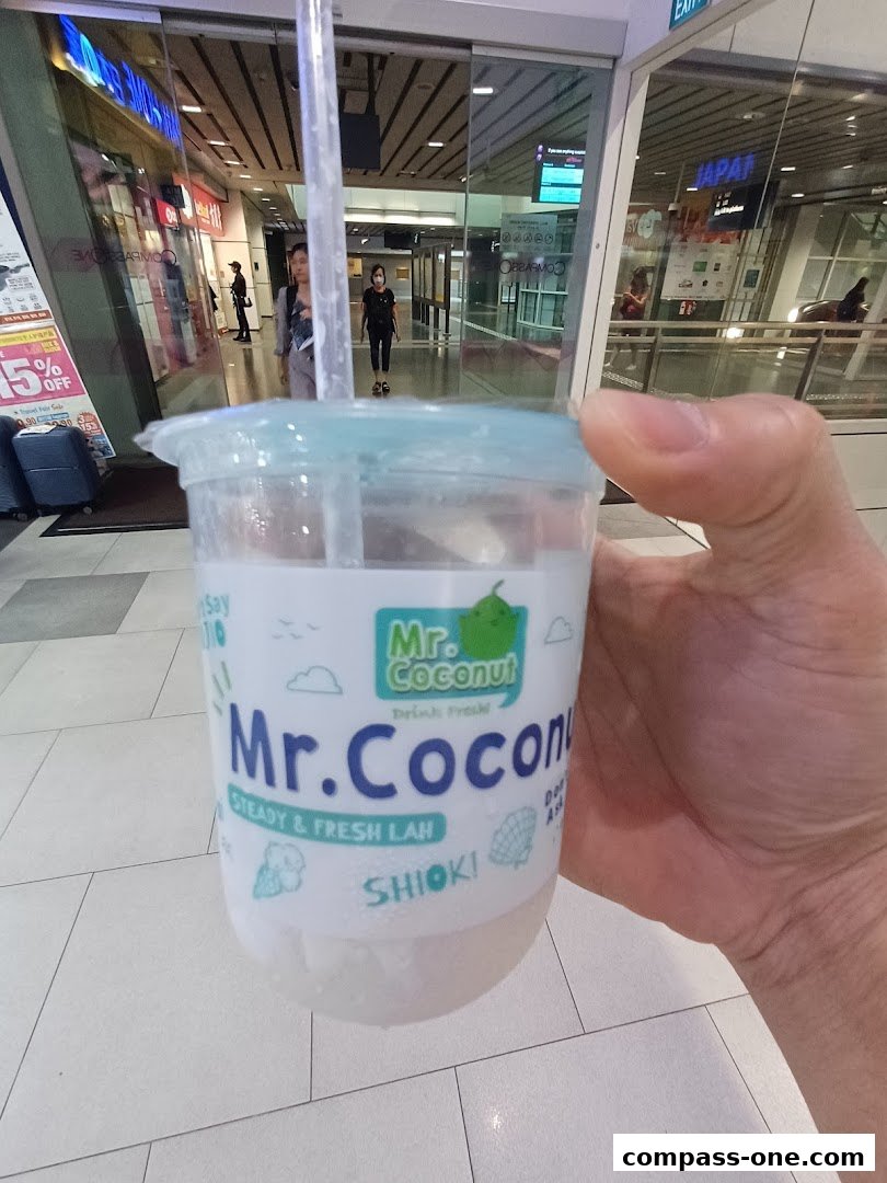 A hand holds a Mr. Coconut drink in front of a shopping mall corridor.