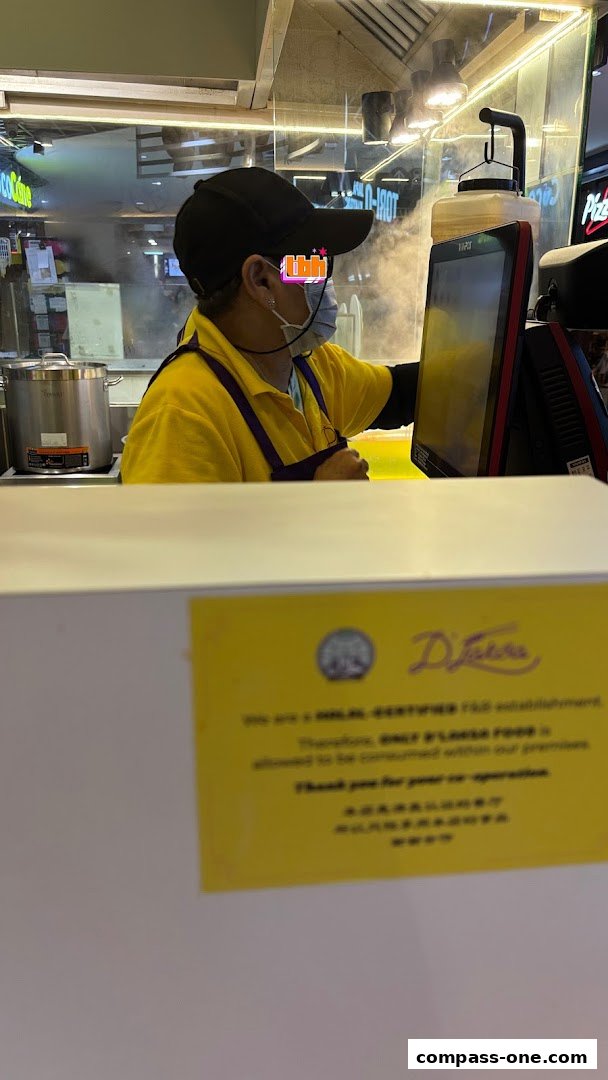 A staff member in a yellow shirt and black cap works behind a counter at D Laksa.