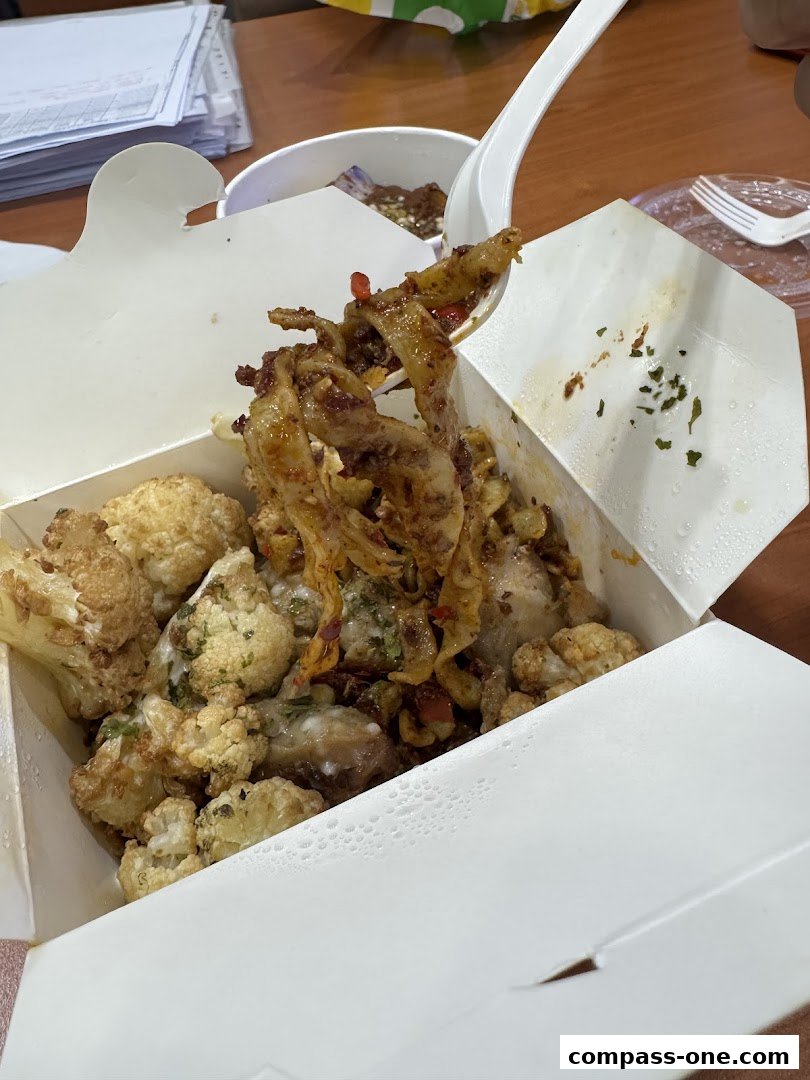 A close-up of a takeout box filled with pasta and cauliflower, with a spoon lifting some pasta.