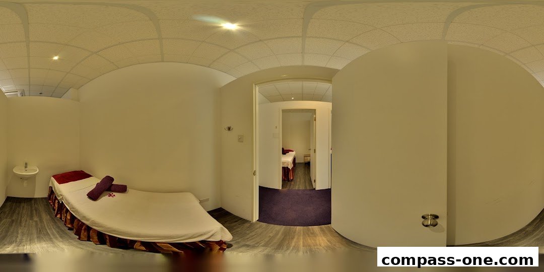 Interior view of a spa room with a massage bed and a sink, and another room visible through a doorway.
