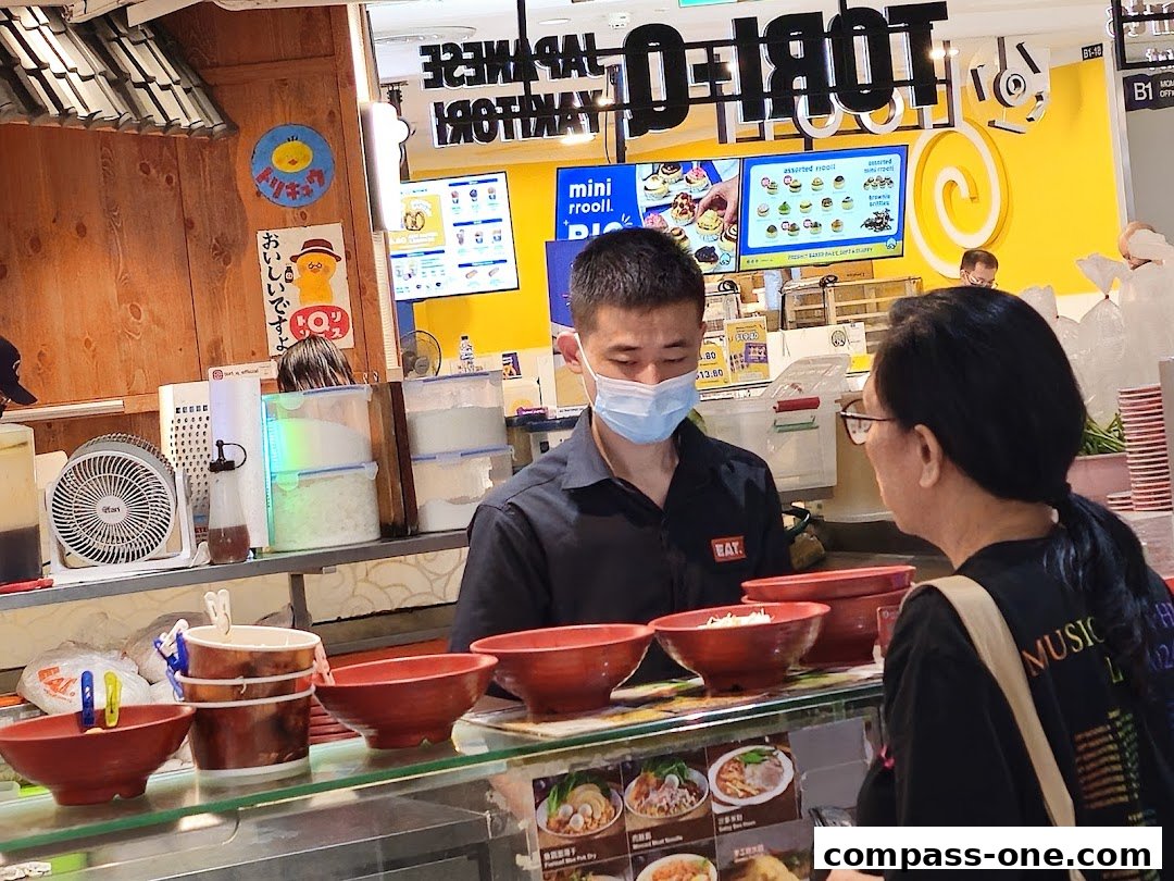 A customer interacts with a staff member at the EAT. food stall, with menu displays in the background.