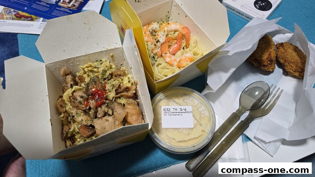 A spread of takeaway pasta dishes, chicken wings, and a side of sauce with cutlery.