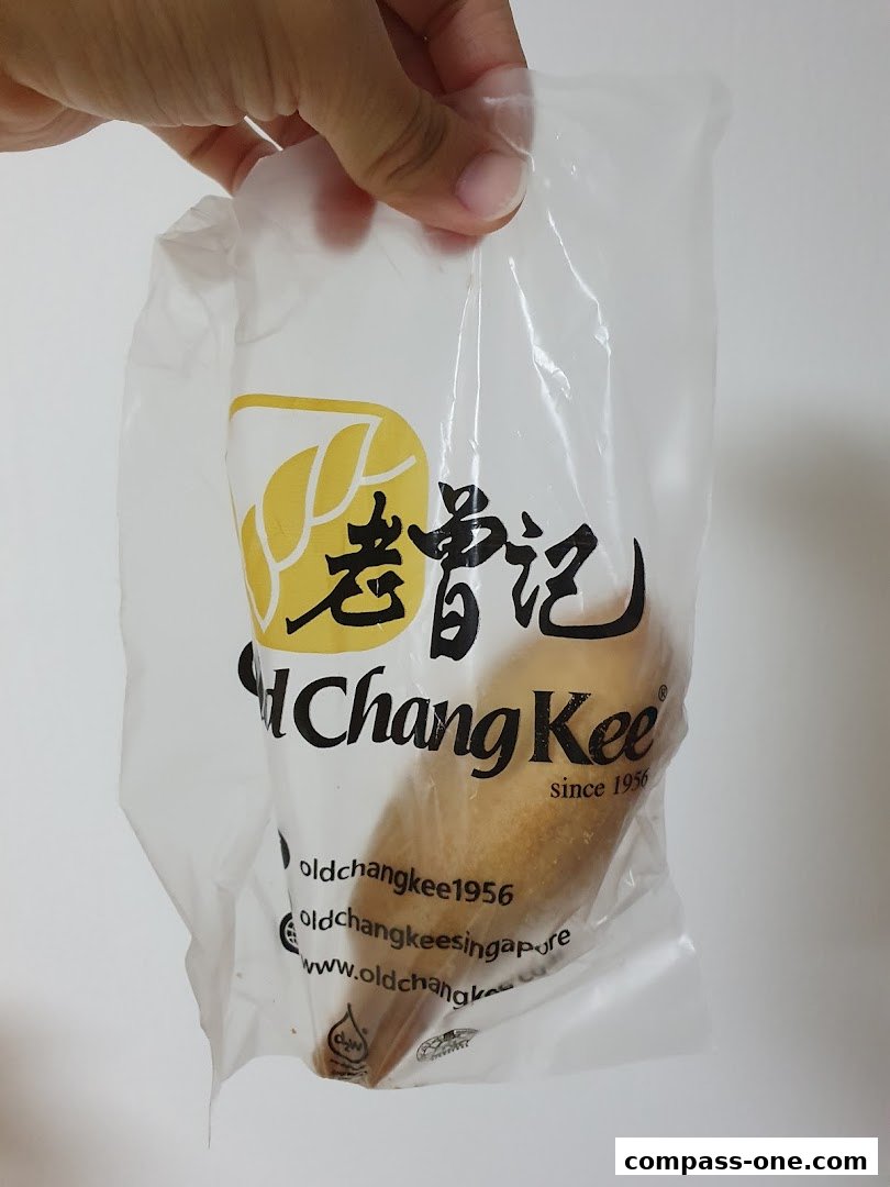 A hand holds a transparent plastic bag with the Old Chang Kee logo and a pastry inside.