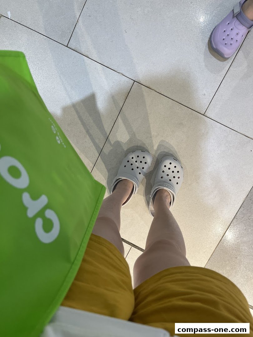A person wearing grey Crocs and yellow shorts, holding a green Crocs shopping bag.