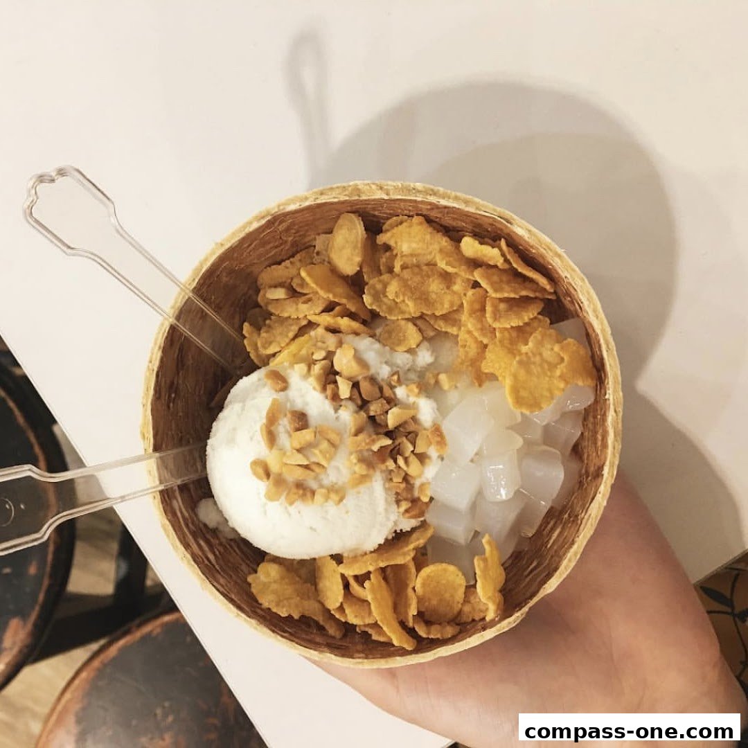 A hand holds a coconut shell filled with ice cream, cornflakes, and diced coconut.