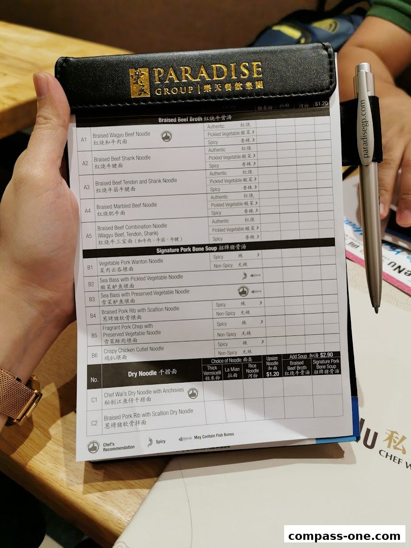 A person holding a menu from LeNu Chefs Wai Noodle Bar, showing various noodle dishes and prices.