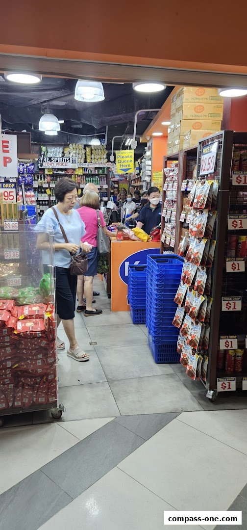 Customers queue at the entrance of a brightly lit convenience store with shelves stocked with goods.